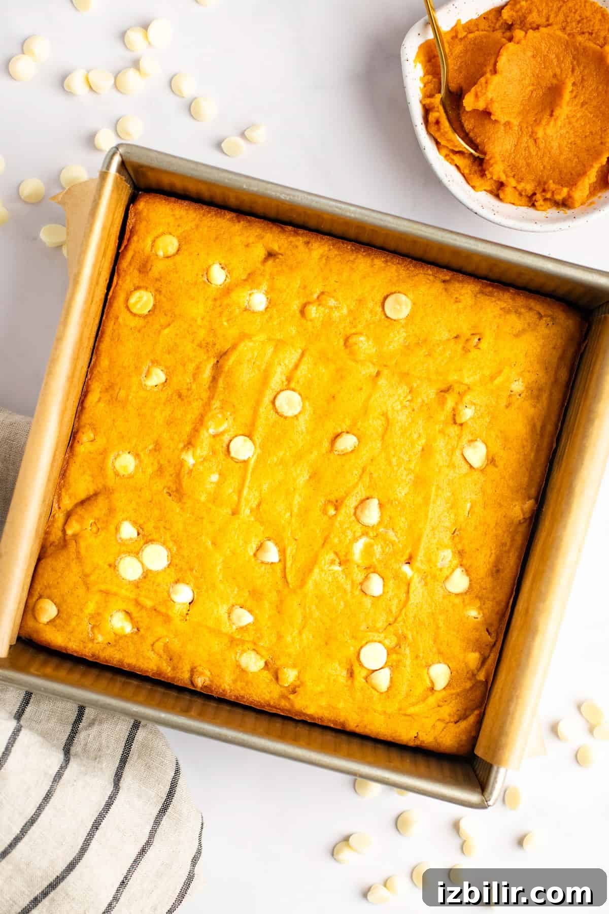 Chewy Pumpkin Blondies Recipe with Spiced Brown Sugar 4 A square pan of orange-colored baked goods with white chocolate chips. A small bowl of pumpkin puree and a spoon are placed beside the pan. A striped cloth and scattered chocolate chips are also visible.