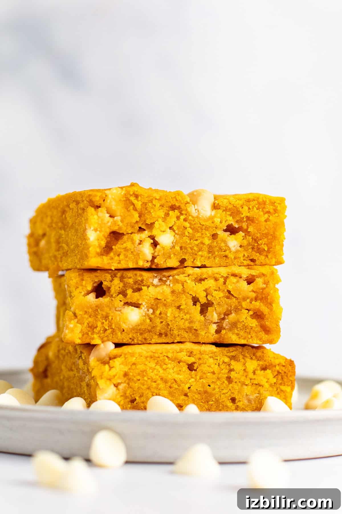 Chewy Pumpkin Blondies Recipe with Spiced Brown Sugar 5 A stack of three pumpkin-flavored blondies with white chocolate chips sits on a grey plate, surrounded by scattered white chocolate chips, against a white background.