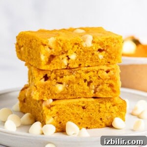 A stack of three pumpkin blondies with white chocolate chips on a white plate. Scattered white chocolate chips surround the stack.