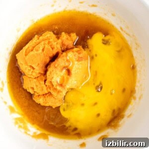 Chewy Pumpkin Blondies Recipe with Spiced Brown Sugar 8 A mixing bowl contains mashed sweet potatoes, yolk-like liquid, and brown syrupy substance being prepared for a recipe.