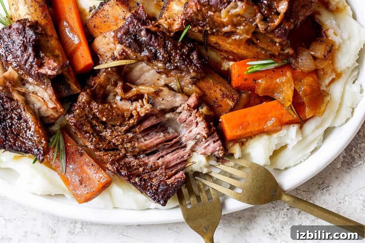 Braised Short Ribs That Fall Off the Bone 2 A plate of tender braised beef short ribs with carrots, garnished with rosemary, served on a bed of creamy mashed potatoes. Two forks are placed beside the dish.