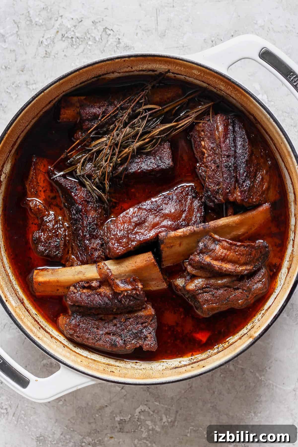 Braised Short Ribs That Fall Off the Bone 4 A large pot filled with braised short ribs in a rich, dark sauce, garnished with herbs on a light gray surface.