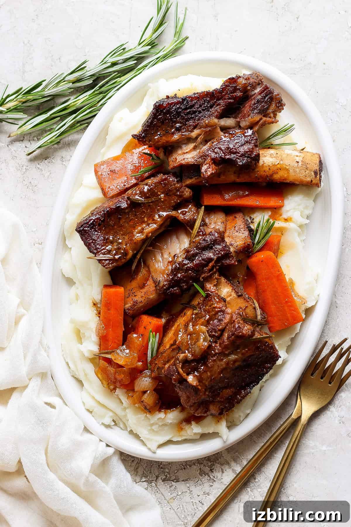 Braised Short Ribs That Fall Off the Bone 6 A white oval plate with mashed potatoes, roasted carrot slices, and beef short ribs garnished with fresh rosemary sprigs. A fork and knife are placed beside the plate on a white surface.