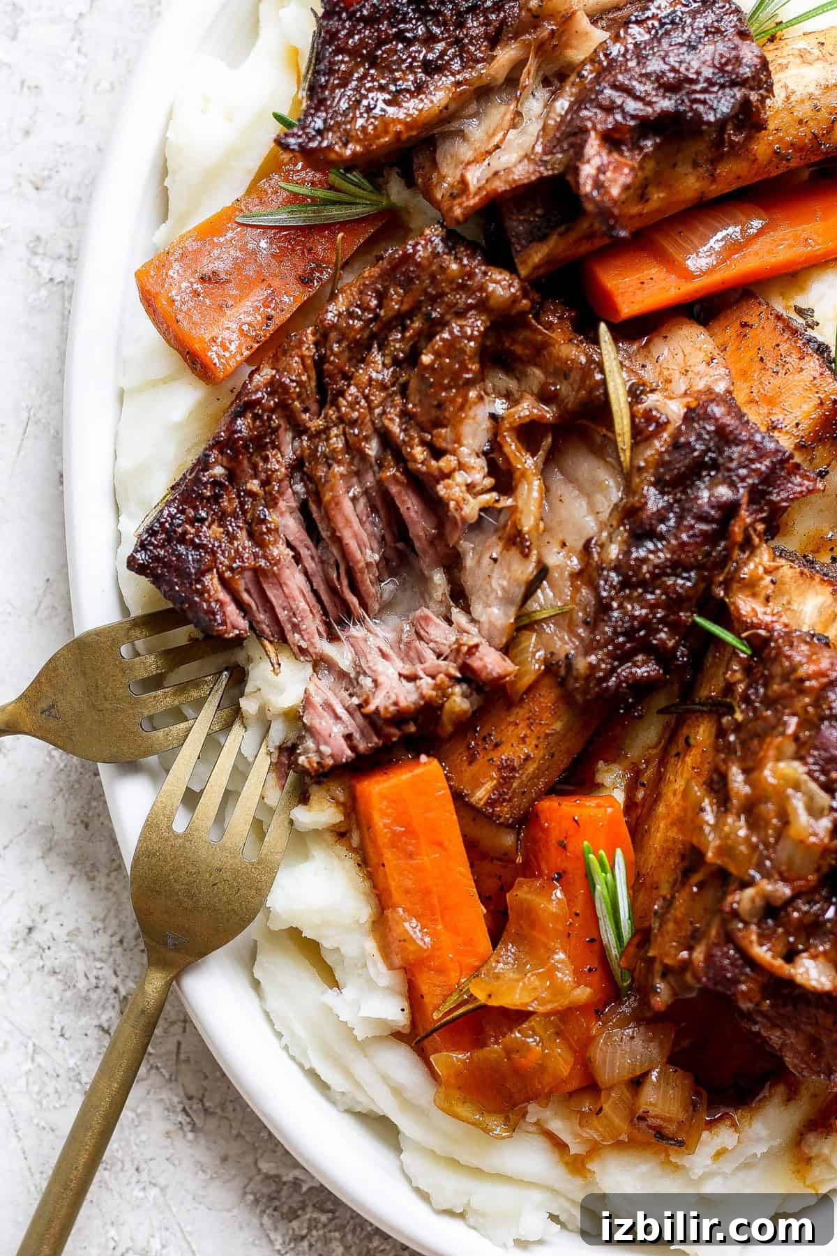 Braised Short Ribs That Fall Off the Bone 7 A plate of tender braised beef short ribs with carrots, garnished with rosemary, served on a bed of creamy mashed potatoes. Two forks are placed beside the dish.