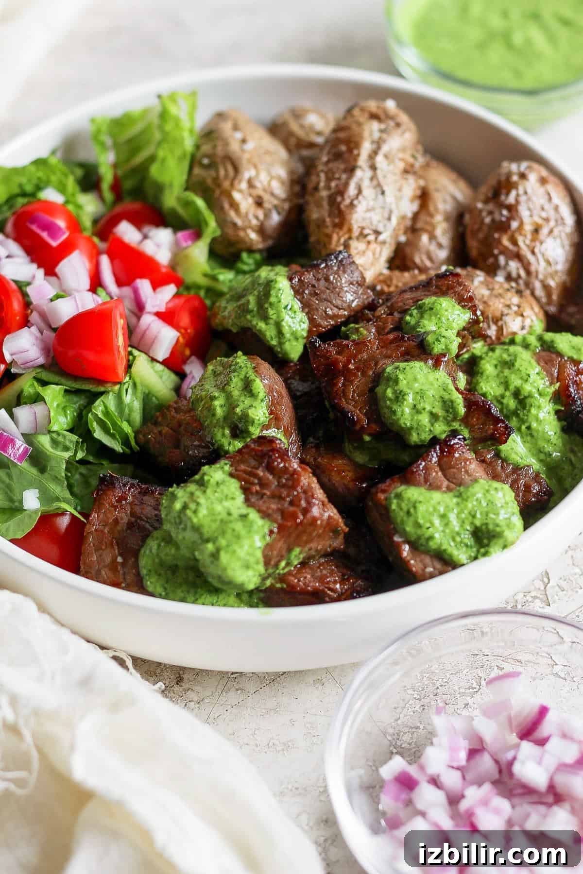 Fit Foodie Cooking Club: September 2024 Seasonal Recipes 5 A bowl with grilled steak topped with green chimi-churri sauce, roasted potatoes, and a salad of lettuce, cherry tomatoes, and red onions. A small dish of chopped red onions is beside the bowl.