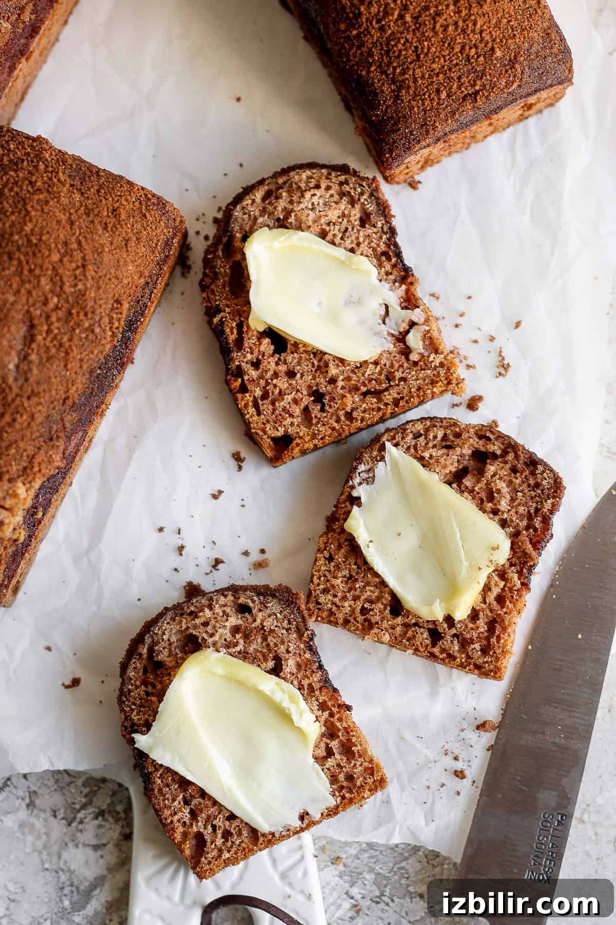 Mini Cinnamon Crunch Loaves Recipe 5 Two slices of dark bread with butter on a parchment paper, next to a knife with a partially visible bread loaf.