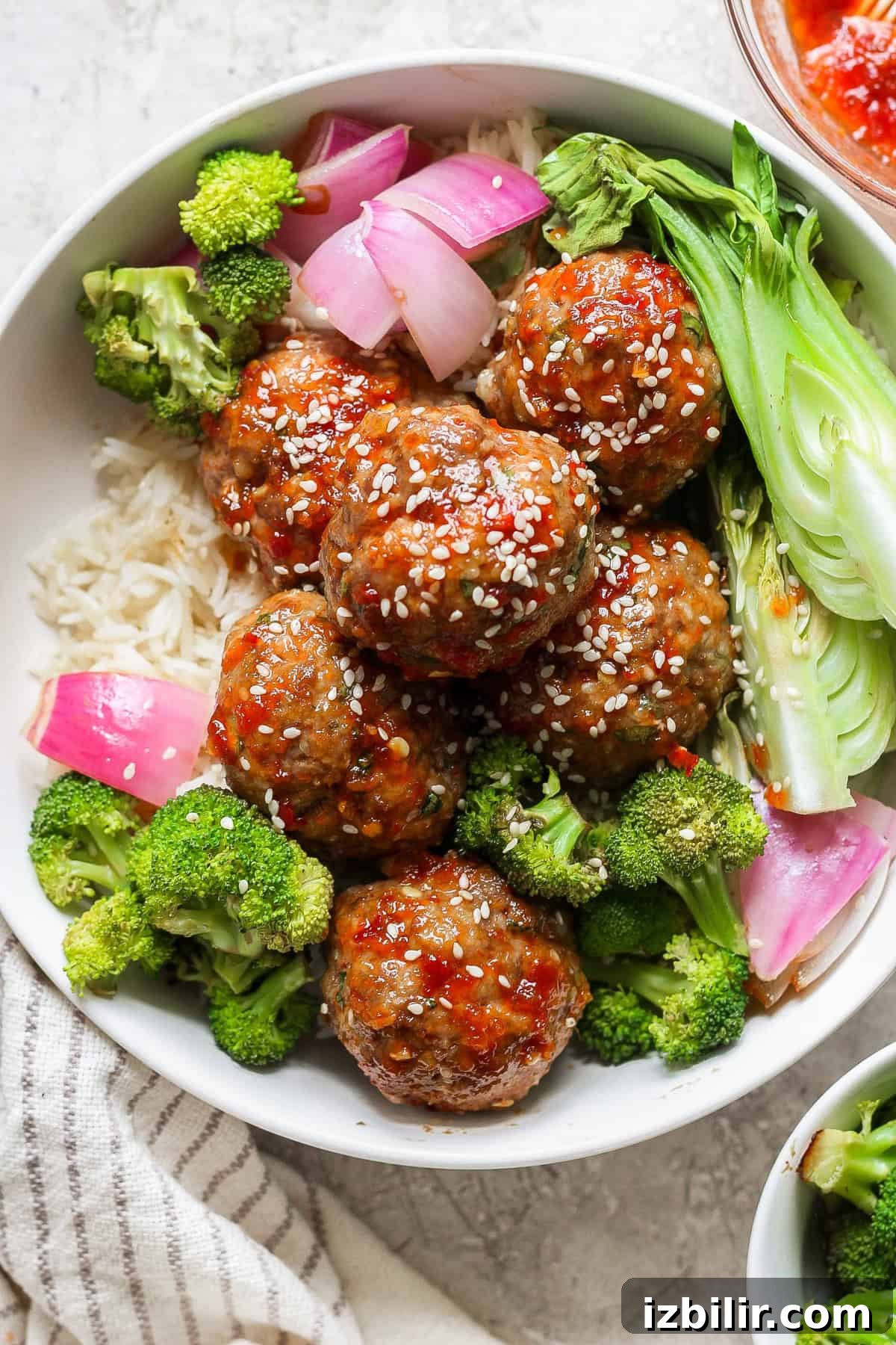 A bowl of rice topped with meatballs glazed in a red sauce, garnished with sesame seeds, and accompanied by broccoli, bok choy, and red onion.