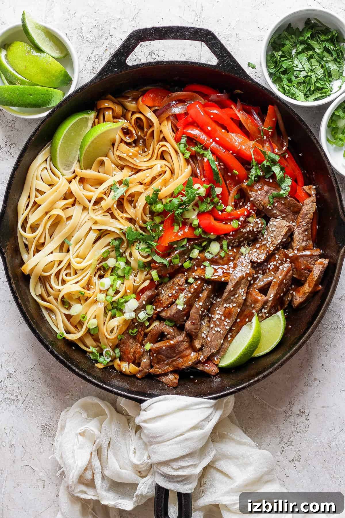 A cast iron skillet contains cooked noodles, sliced beef, red bell peppers, and lime wedges.