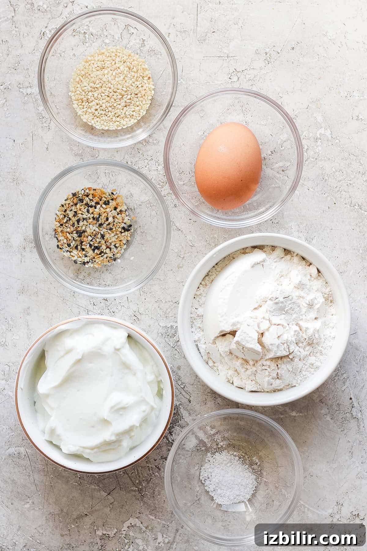 Six bowls containing various ingredients and one egg on a textured surface. The bowls hold sesame seeds, a spice blend, flour, a creamy substance, and salt.