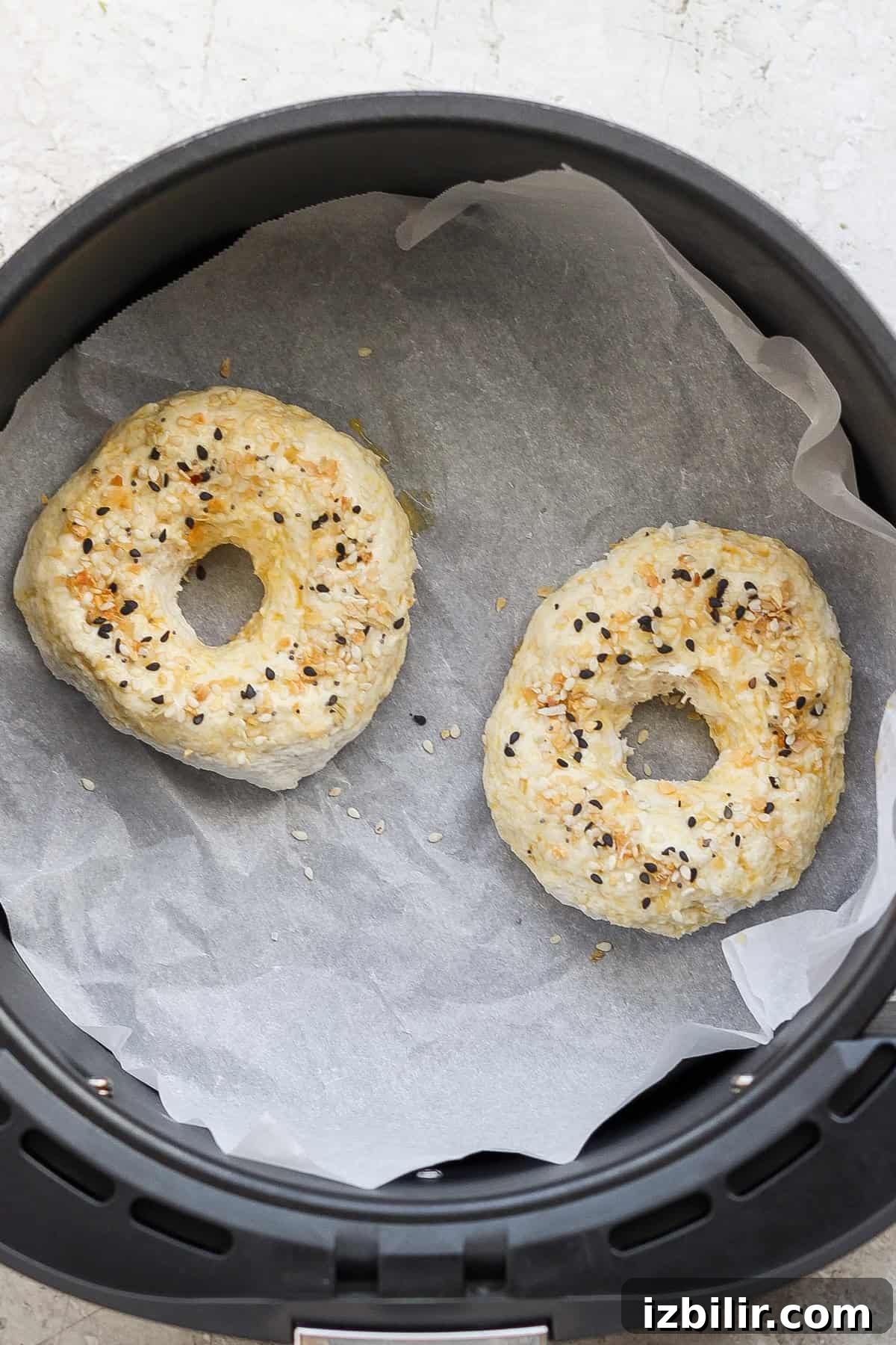 Two bagels with seasoning on top are being cooked on parchment paper inside an air fryer.