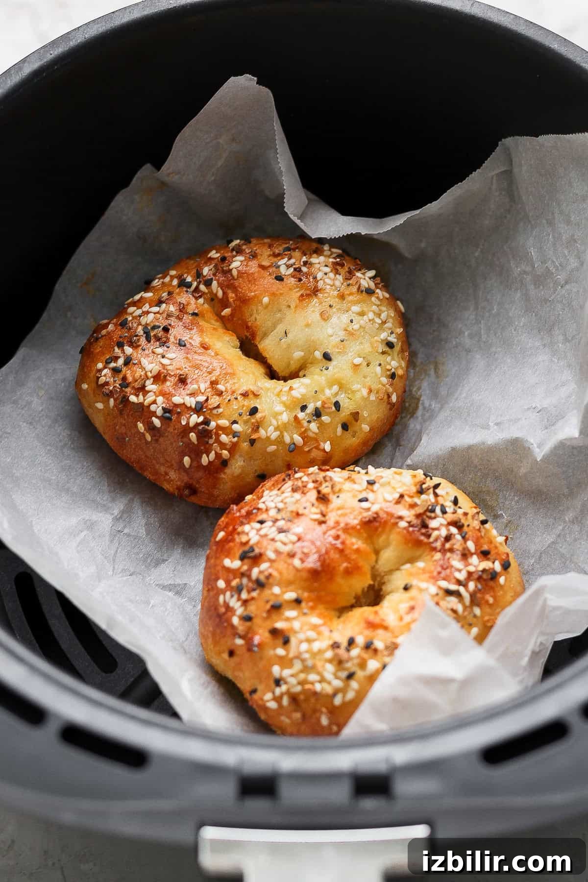 Two bagels topped with sesame and poppy seeds are baking on parchment paper inside an air fryer basket.
