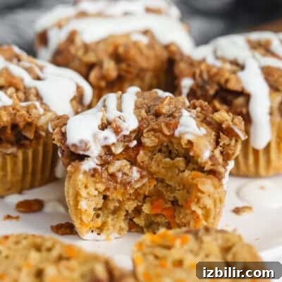 Close-up of a carrot muffin with a crumbly topping and white icing. One muffin has a bite taken out, revealing a moist interior with visible carrot pieces.