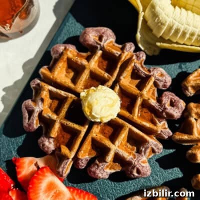 A waffle topped with a dollop of butter is surrounded by sliced bananas and strawberries on a dark surface.