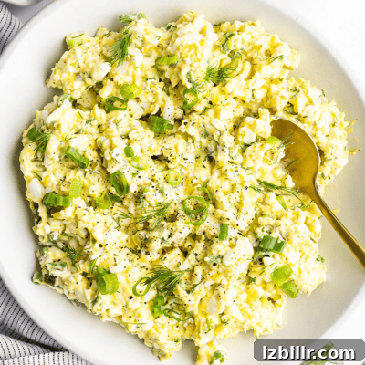 A bowl of egg salad with chopped green onions and dill, accompanied by a gold spoon.