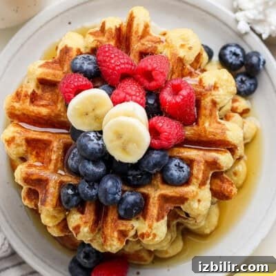 A stack of waffles topped with blueberries, raspberries, banana slices, and drizzled with syrup on a round white plate.