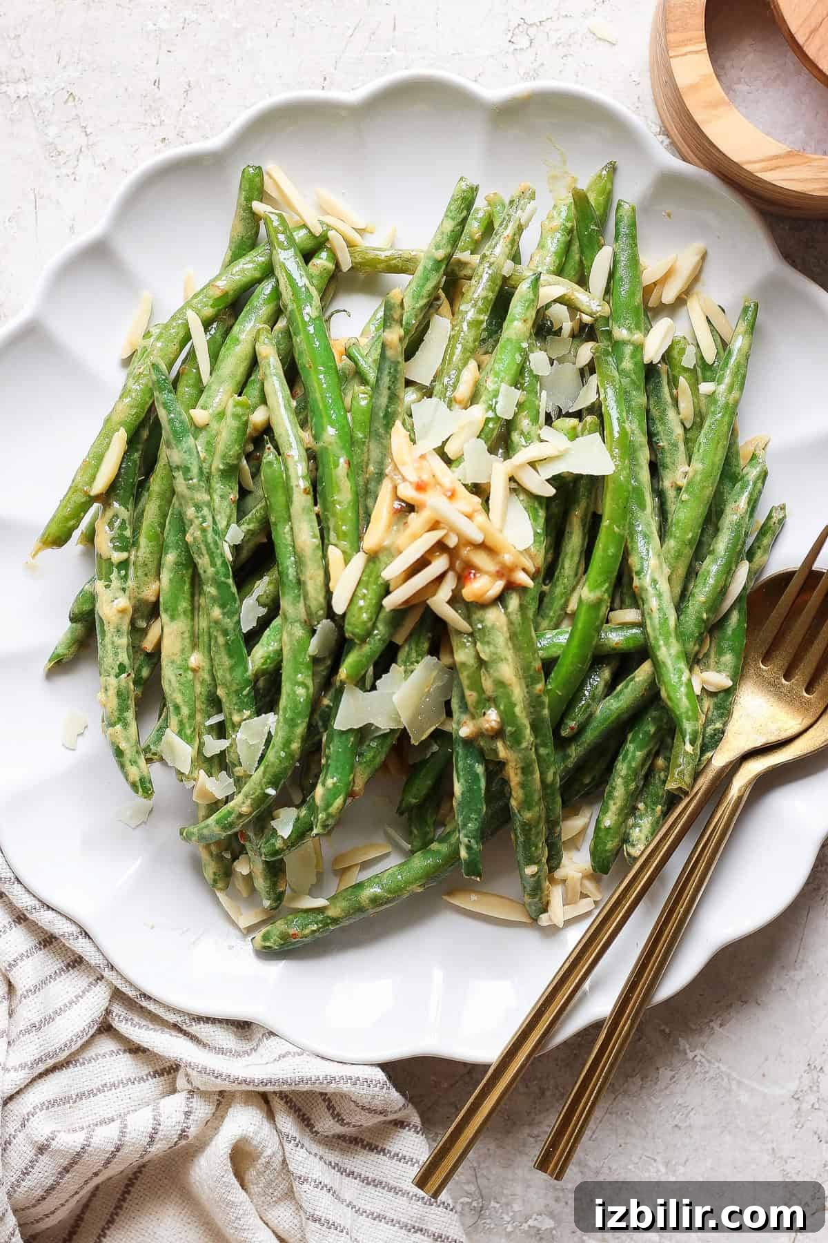 Grandma Ginny's Green Bean Sauté Recipe 2 A plate of green beans topped with sliced almonds and shaved parmesan cheese, served with a gold fork and knife on a white tablecloth.