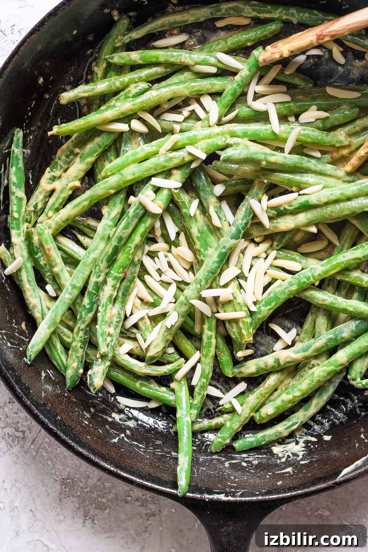 Grandma Ginny's Green Bean Sauté Recipe 6 A skillet filled with green beans coated in a creamy sauce, topped with slivered almonds. A wooden spoon is visible in the top right corner.