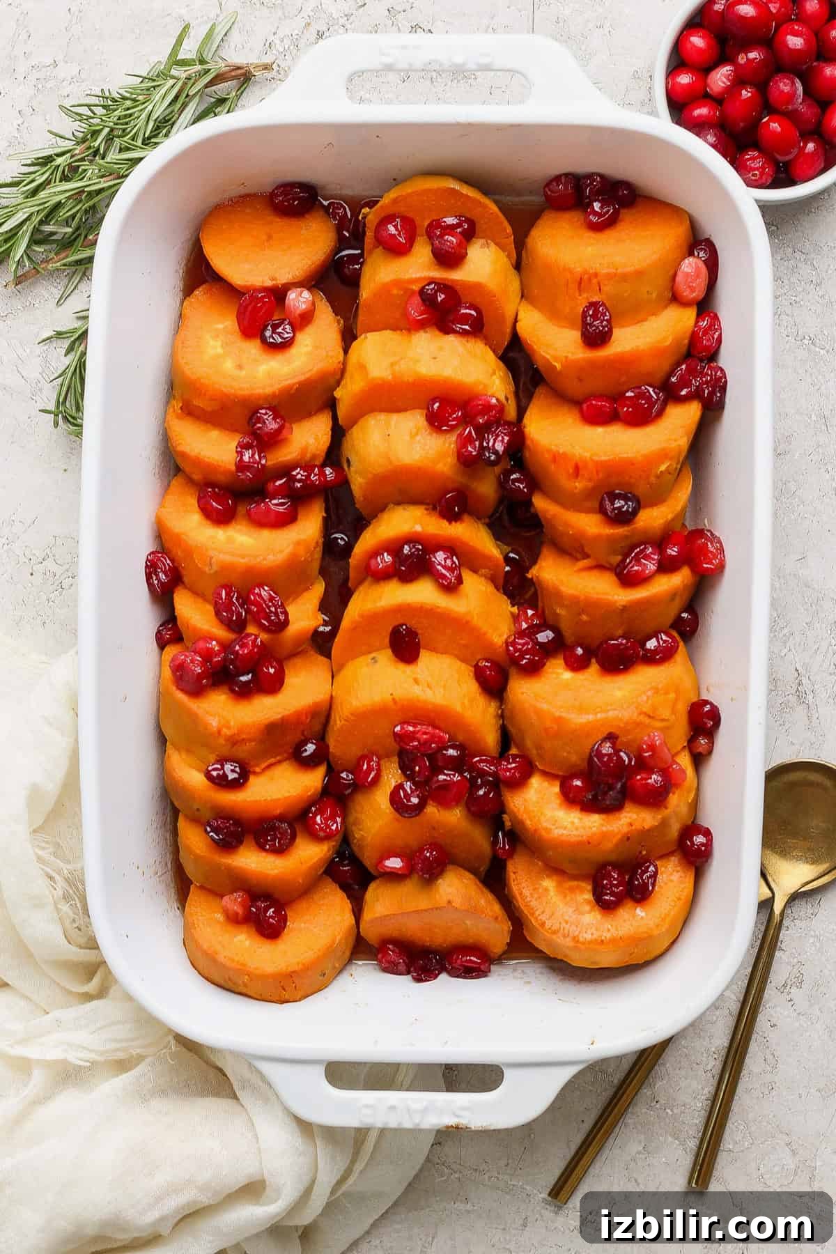 Grandpa Dick's Maple Cranberry Sweet Potatoes Recipe 7 A white baking dish filled with sliced sweet potatoes topped with cranberries. A sprig of rosemary, a bowl of cranberries, and two gold utensils are placed beside the dish.