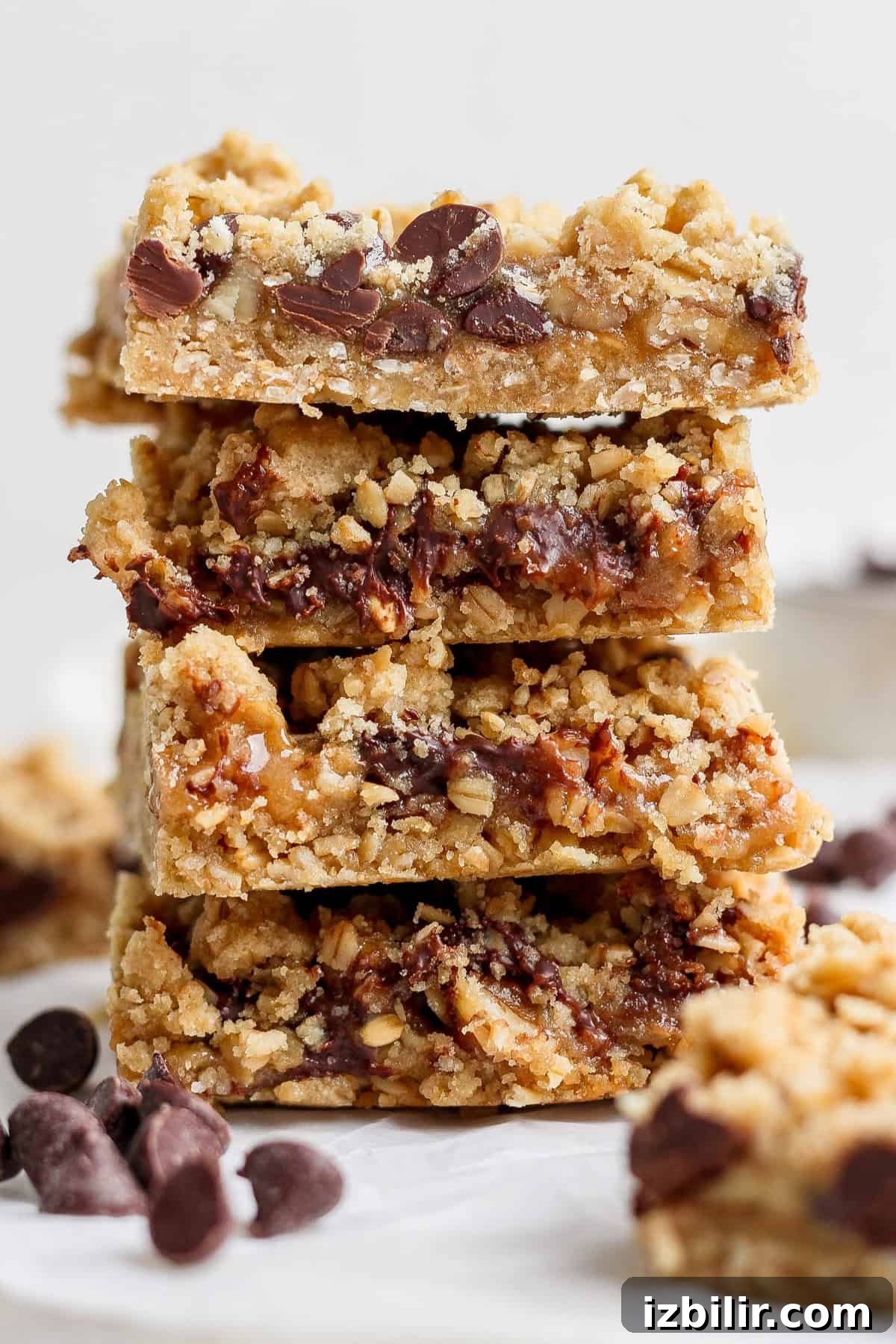 A stack of four chocolate chip oatmeal bars placed on a white surface, with scattered chocolate chips around them.