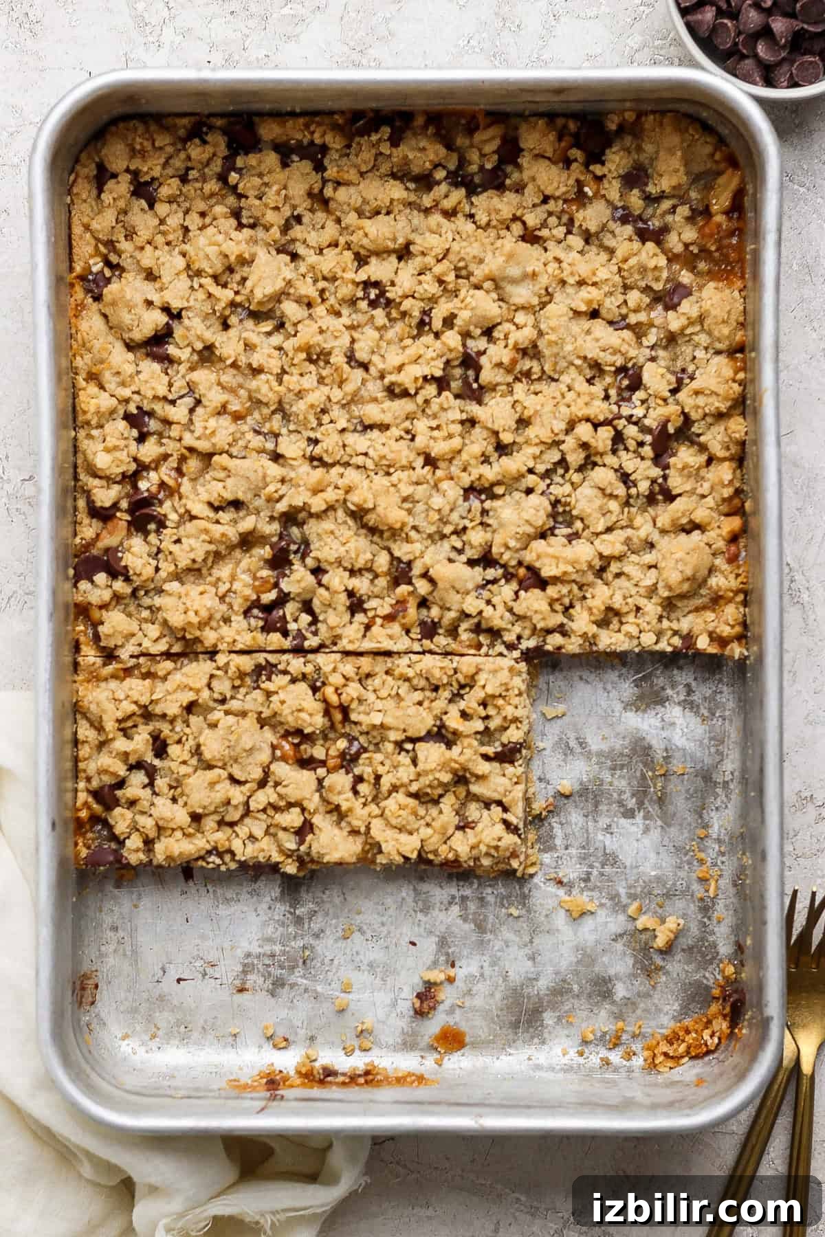 A metal baking pan filled with crumbly dessert bars. Two bars have been cut out, revealing a layered filling, while the rest of the dessert remains uncut. A fork and a napkin are nearby.