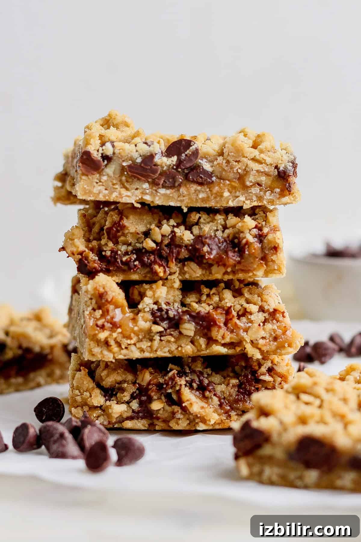 A stack of four chocolate chip oatmeal bars with crumbly tops is displayed on a white surface, surrounded by a few scattered chocolate chips.