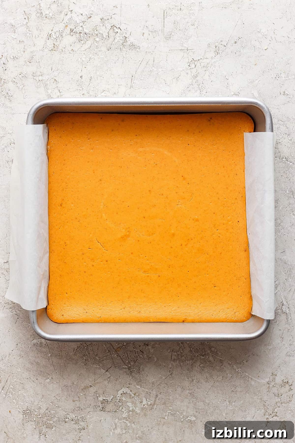 A square pan with a light golden-brown baked cake lined with parchment paper on a textured, light-colored surface.