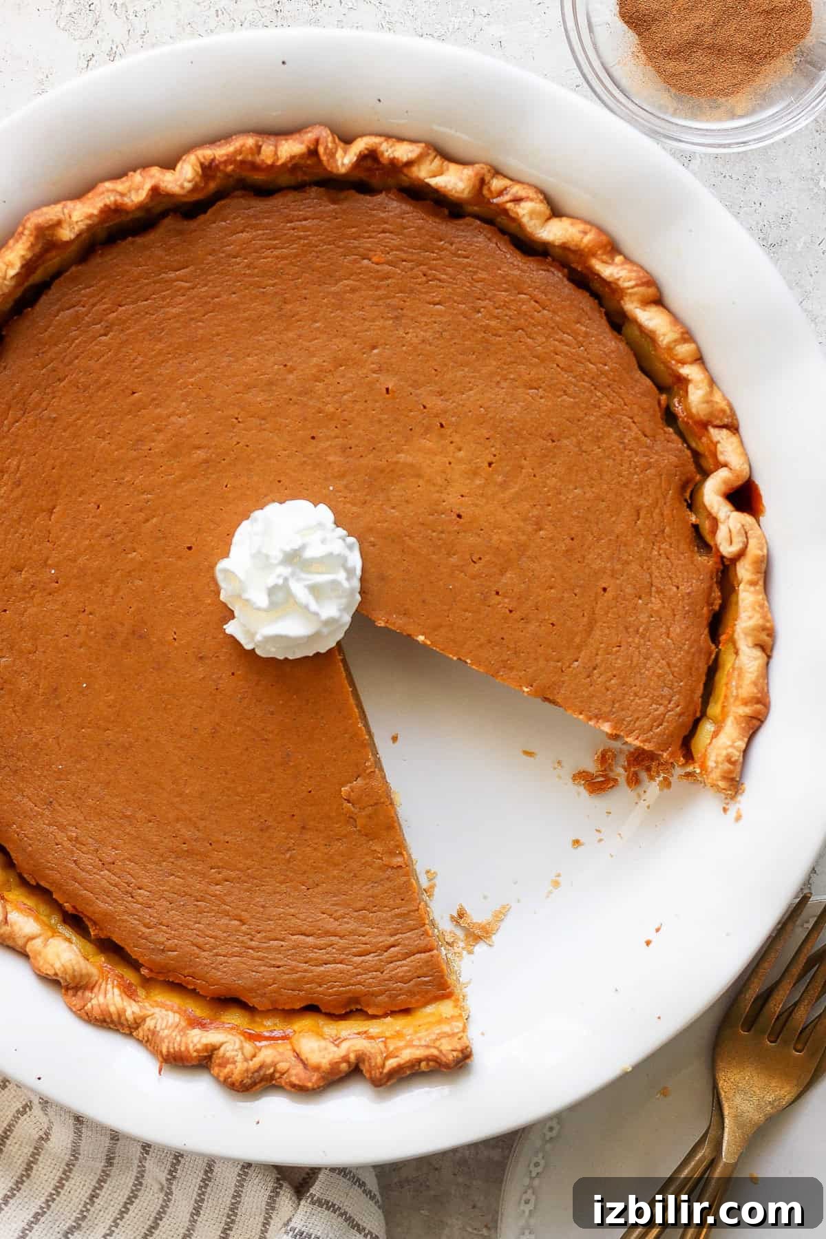 A pumpkin pie with a slice missing, topped with a dollop of whipped cream, in a round white pie dish. There is a folded towel and two gold forks next to the dish.