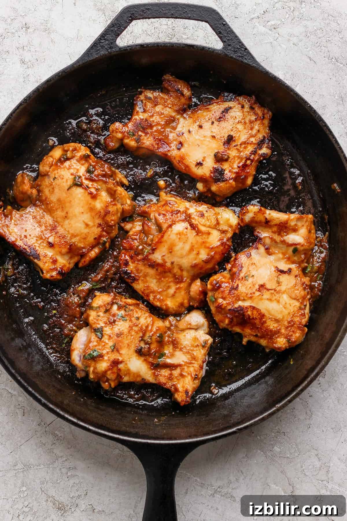 A cast-iron skillet contains five pieces of cooked, seasoned chicken, set on a light-colored surface.