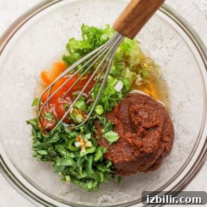 A glass bowl containing chopped green herbs, red sauce, a thick brown paste, and a whisk with a wooden handle on a light-colored surface.