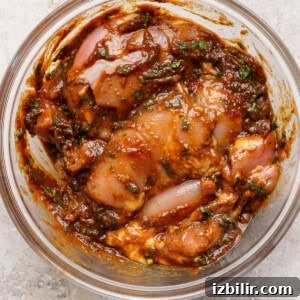 A clear glass bowl containing marinated pieces of raw chicken mixed with herbs and spices in a thick, reddish-brown sauce.