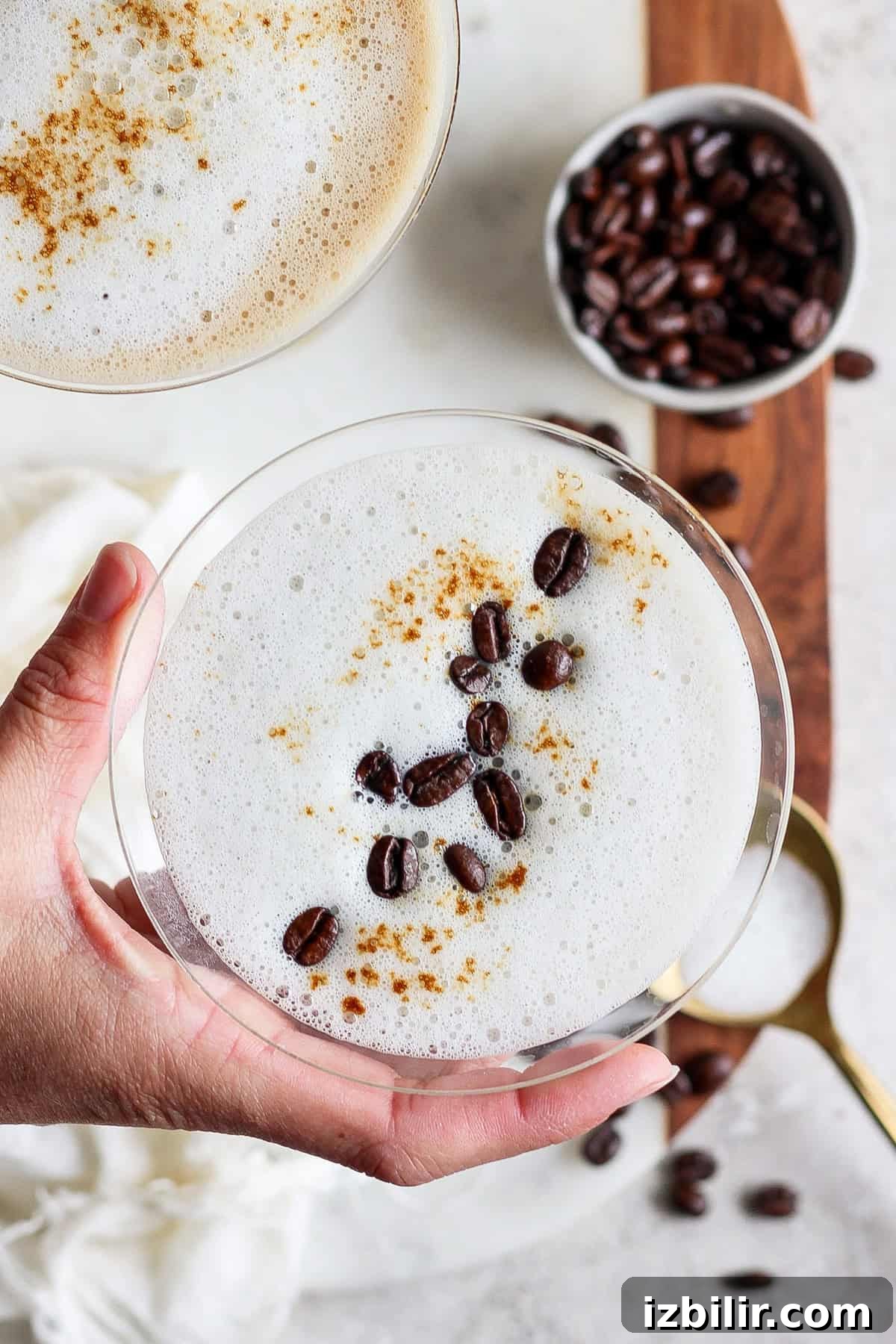 Brown Sugar Shaken Espresso Martini Recipe 5 A hand holding a frothy coffee drink in a glass, garnished with coffee beans and cinnamon