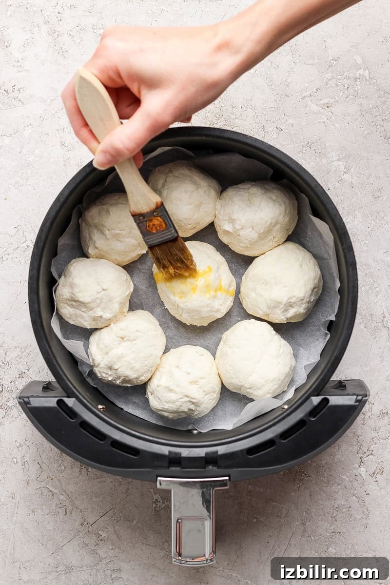 A hand brushes egg wash on dough balls arranged in an air fryer basket lined with parchment paper.