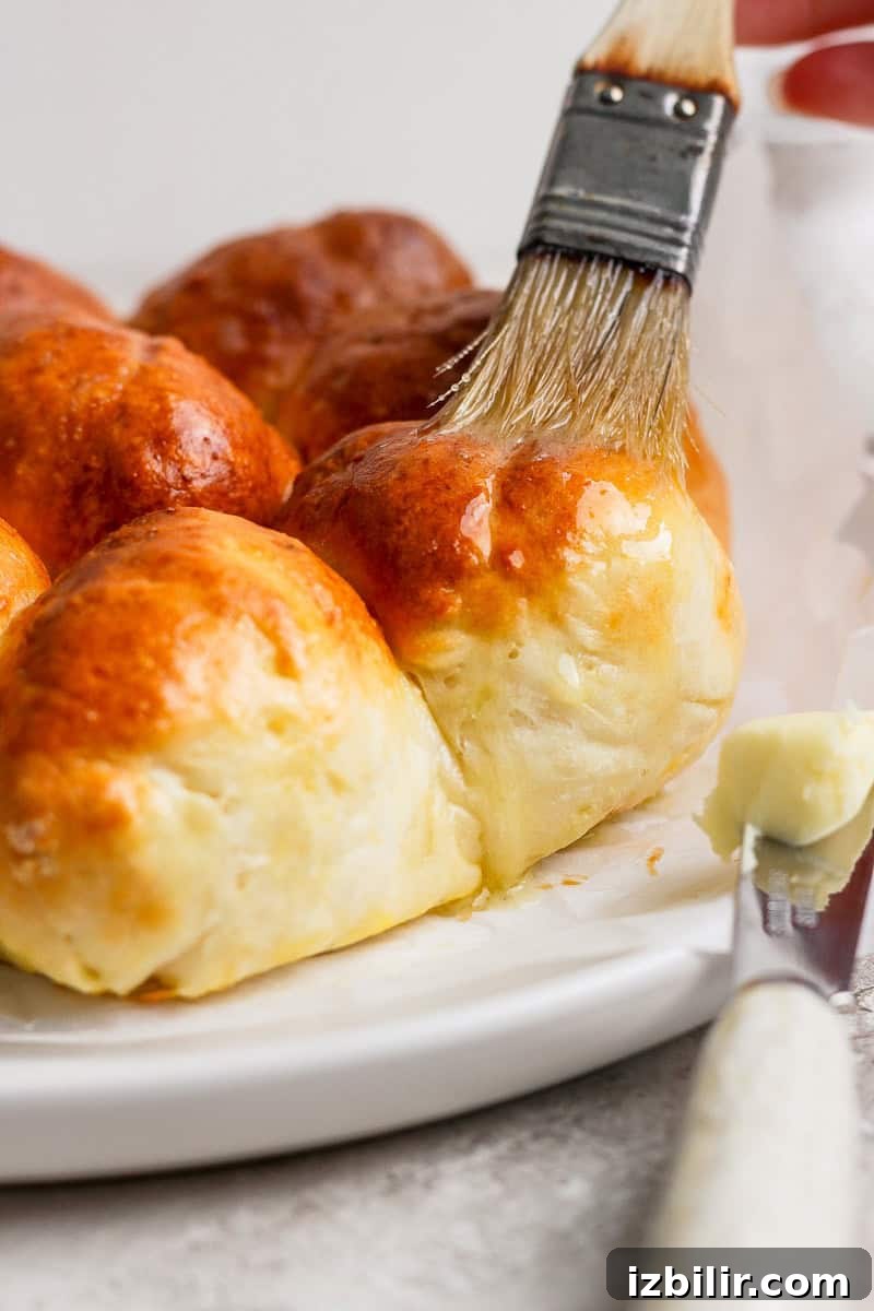 A pastry brush applies butter to freshly baked golden-brown bread rolls on a white plate.