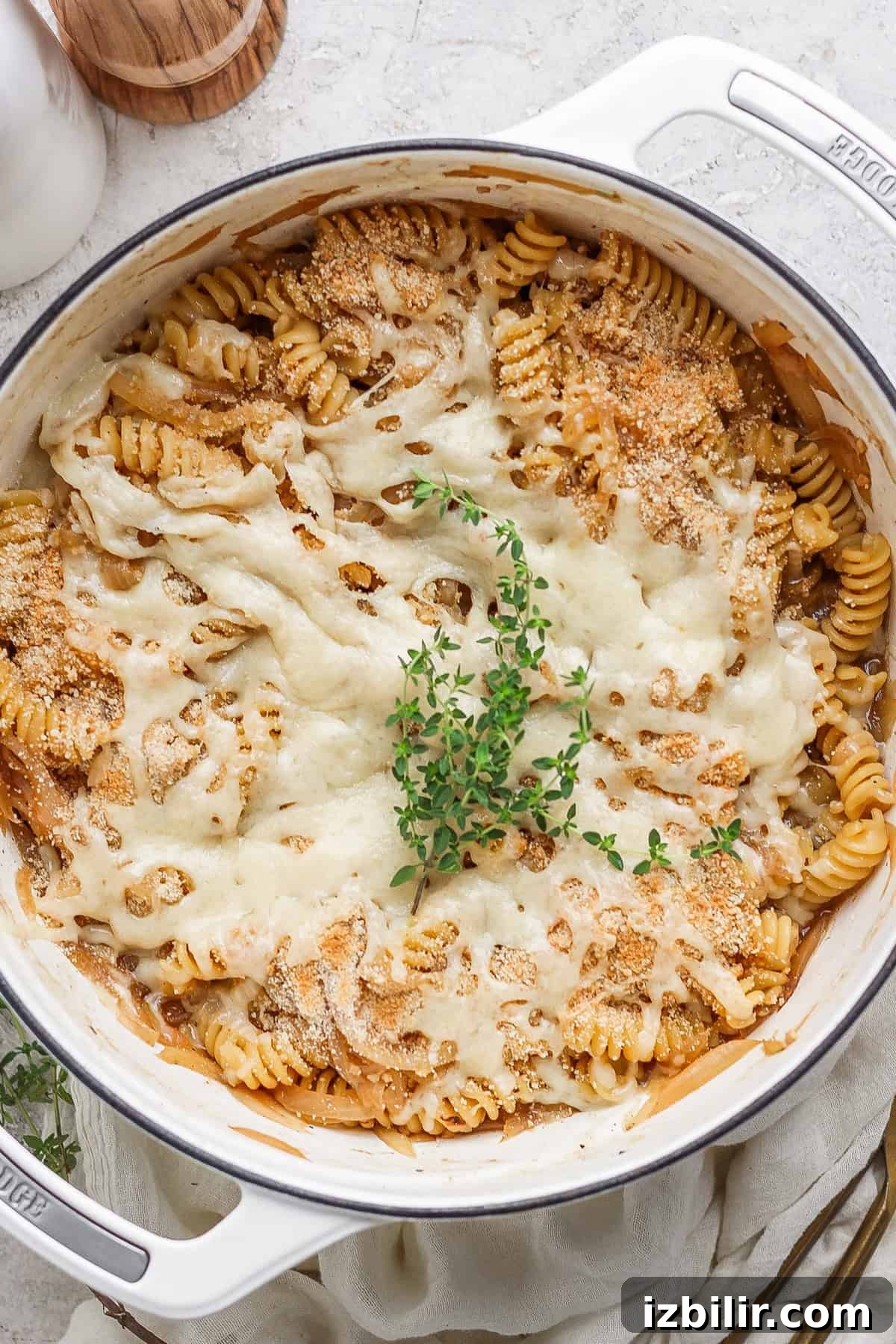 A baked pasta dish in a white pot, featuring rotini pasta covered with melted cheese and garnished with a sprig of fresh herbs on top.