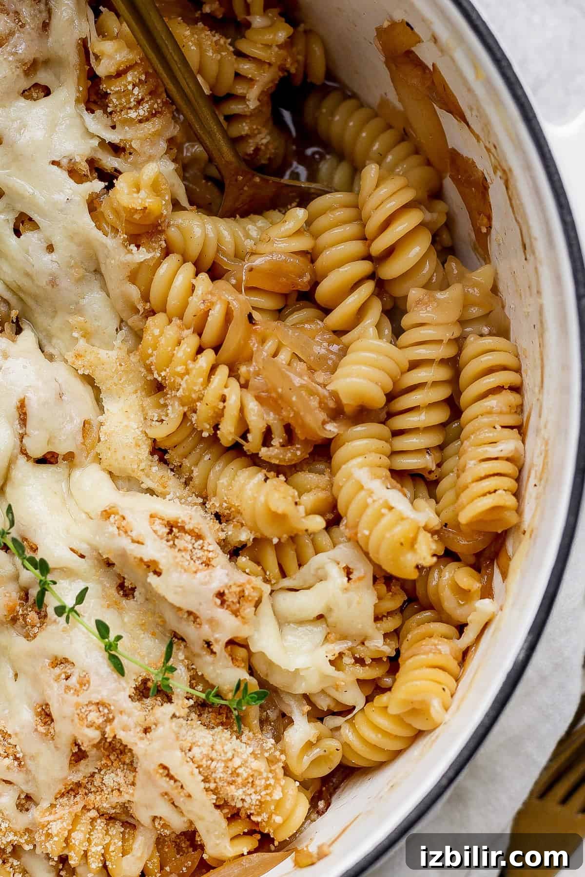 Close-up of baked rotini pasta with melted cheese, breadcrumbs, and herbs in a round dish. A fork is partially visible in the pasta.
