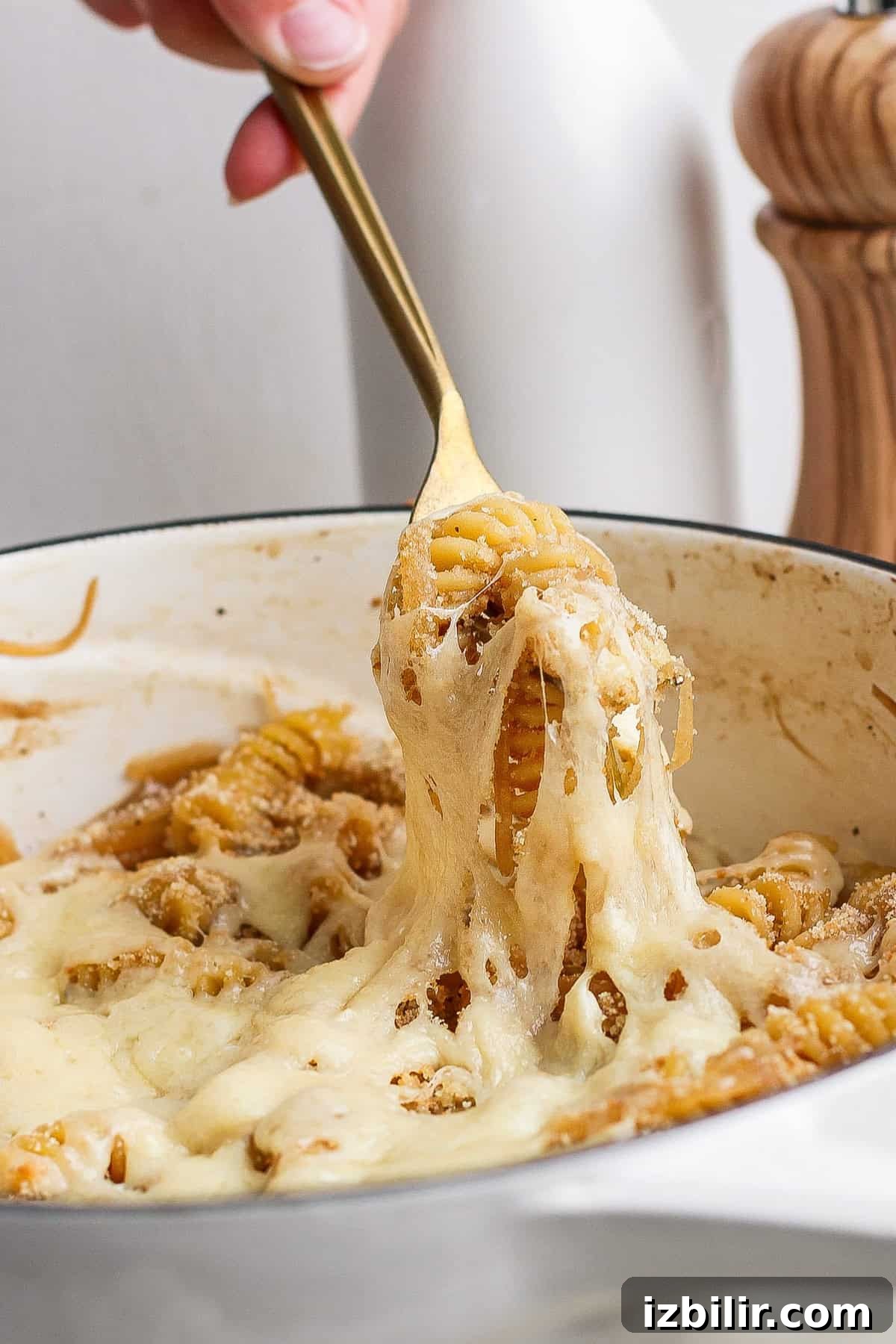 A hand using a golden fork to lift cheesy pasta from a white pot, with melted cheese stretching from the pot.