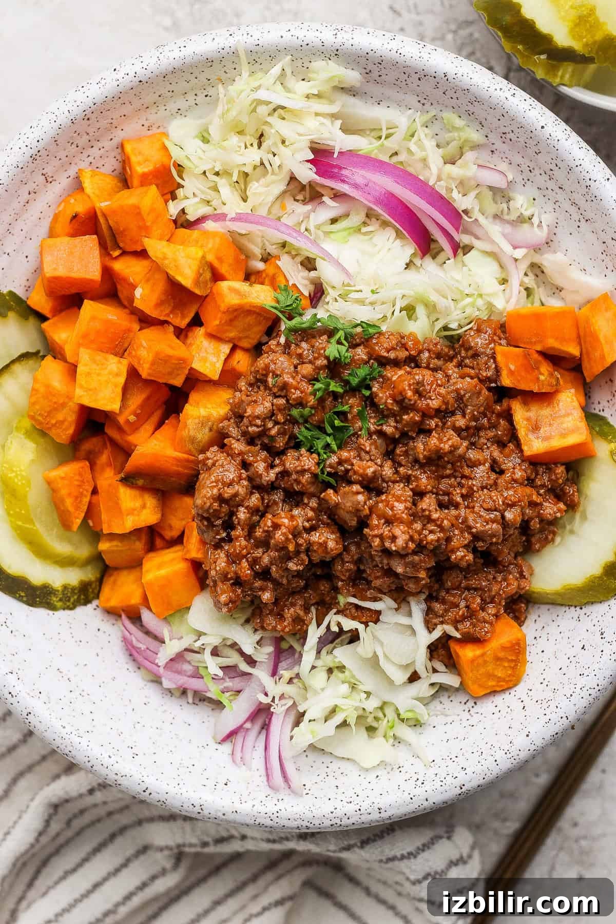 A bowl with ground meat, diced sweet potatoes, sliced pickles, shredded cabbage, and red onion.