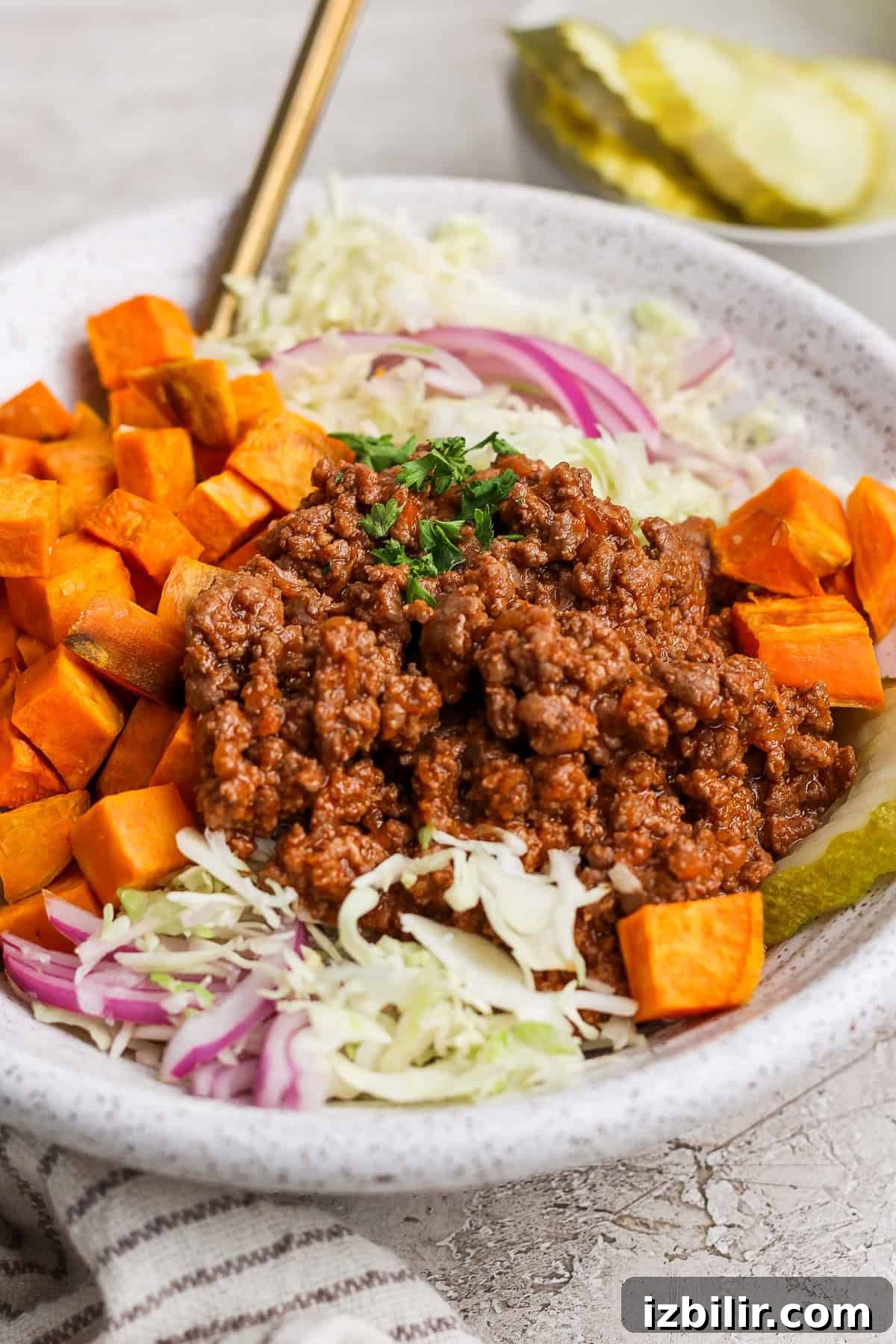 A bowl of salad with shredded cabbage, red onion, diced sweet potatoes, a scoop of seasoned ground meat, and garnished with parsley. Pickle slices are visible in the background.