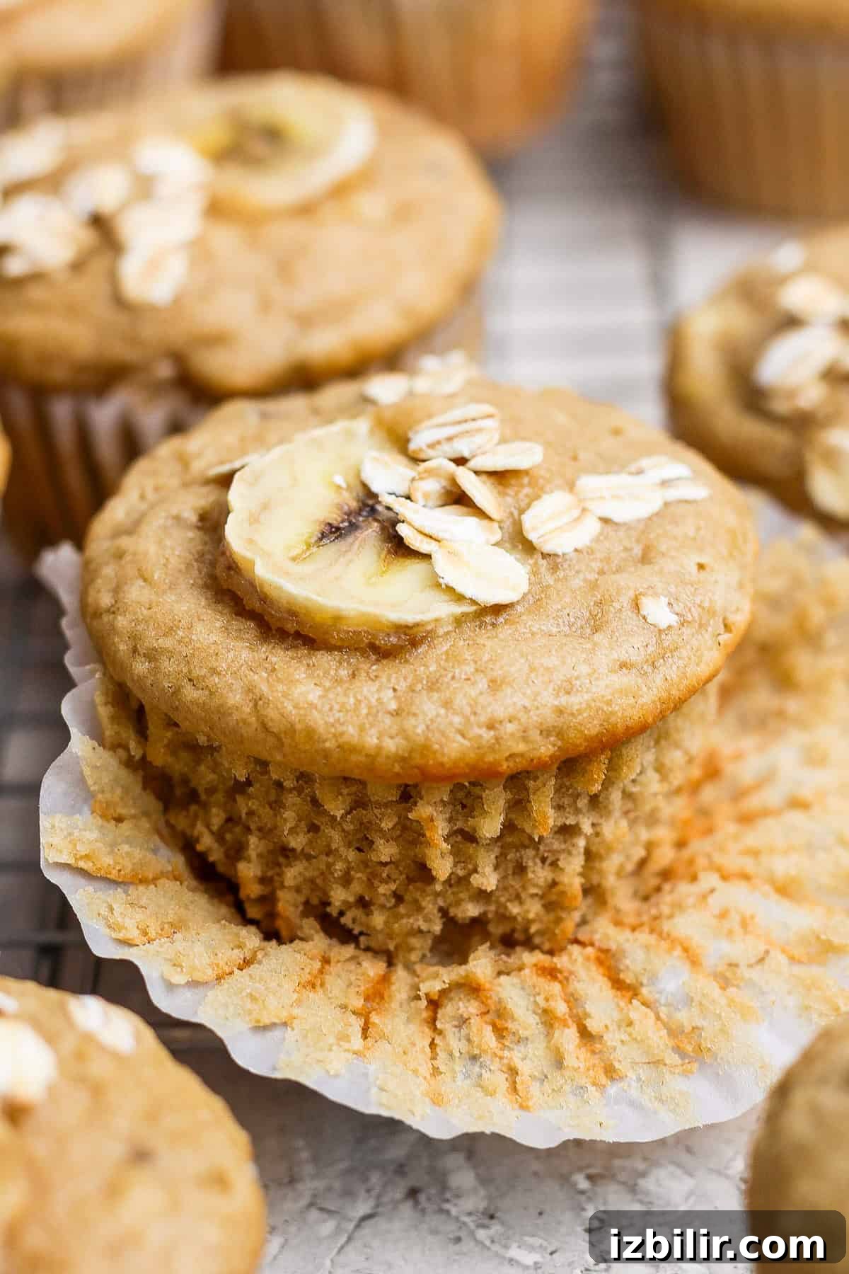 A banana muffin topped with a banana slice and oats, partially unwrapped on a wire rack.