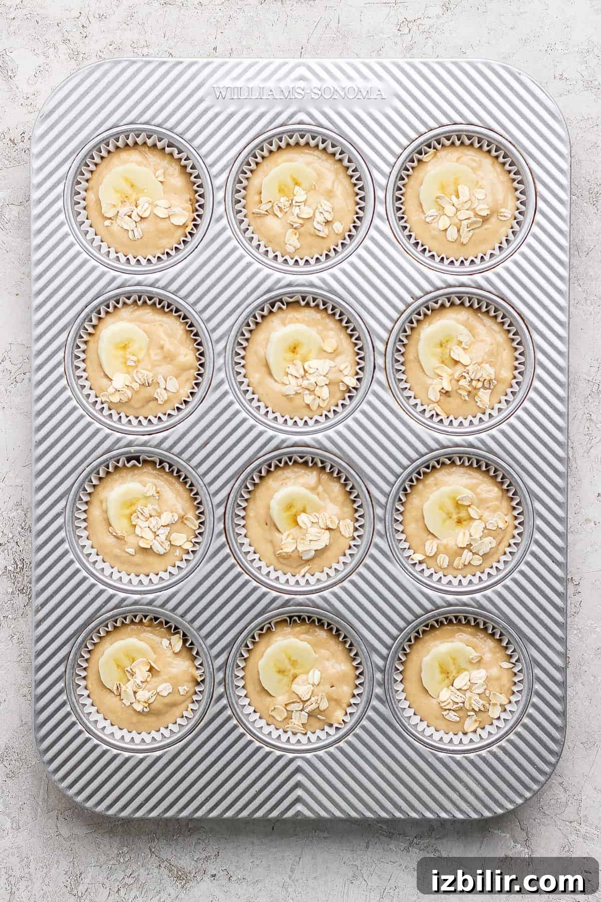 A muffin tray with twelve muffin batter portions, each topped with banana slices and oats, ready for baking.