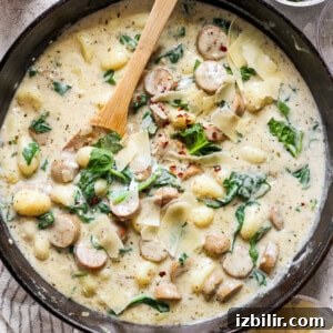 A skillet filled with creamy gnocchi, spinach, and sausage topped with shredded cheese and red pepper flakes. A wooden spoon rests inside the skillet.