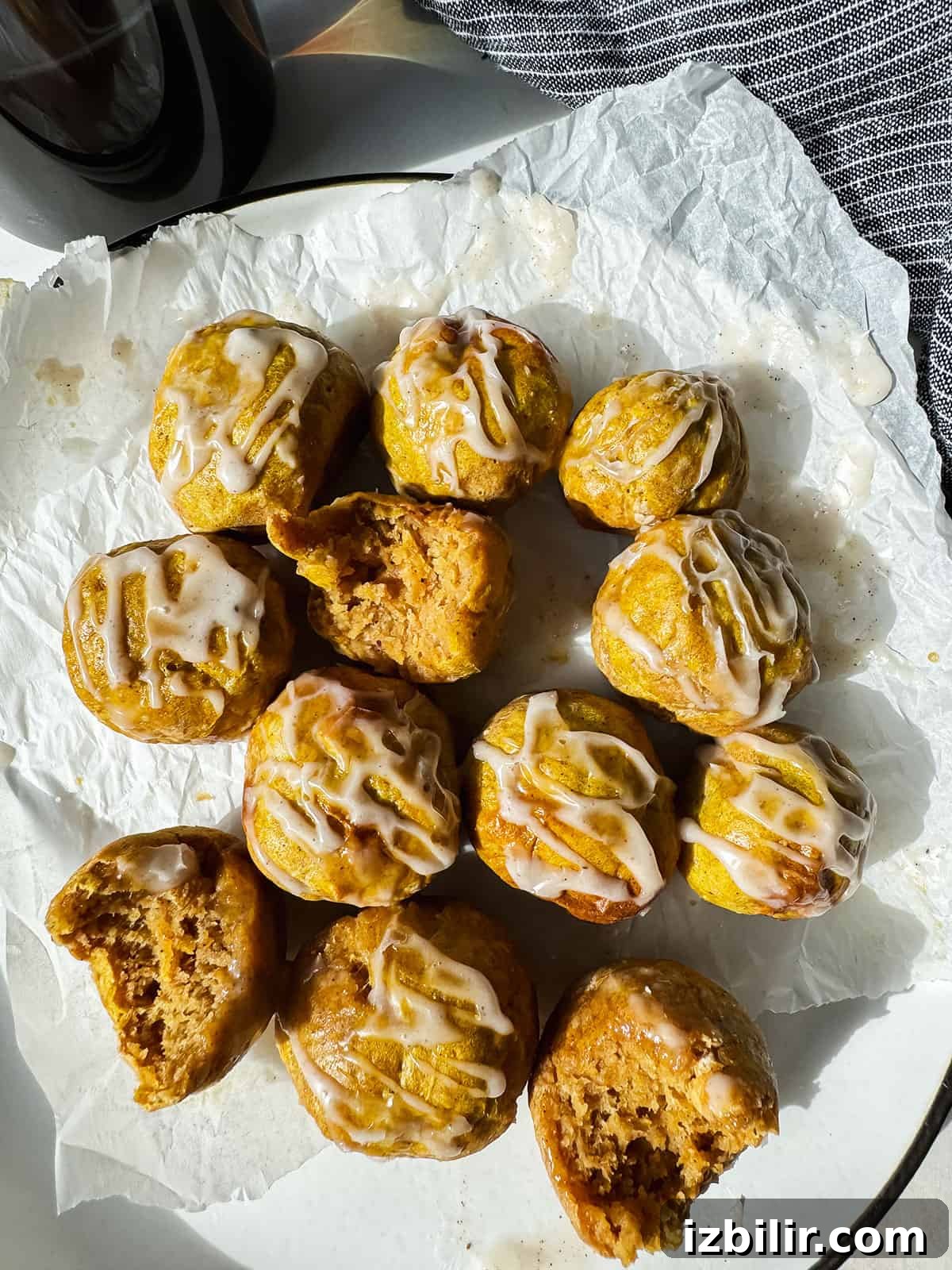 Crispy Air Fryer Pumpkin Bagel Bites Recipe 2 A plate of small, round pastries drizzled with white icing, some partially eaten, on a piece of parchment paper.