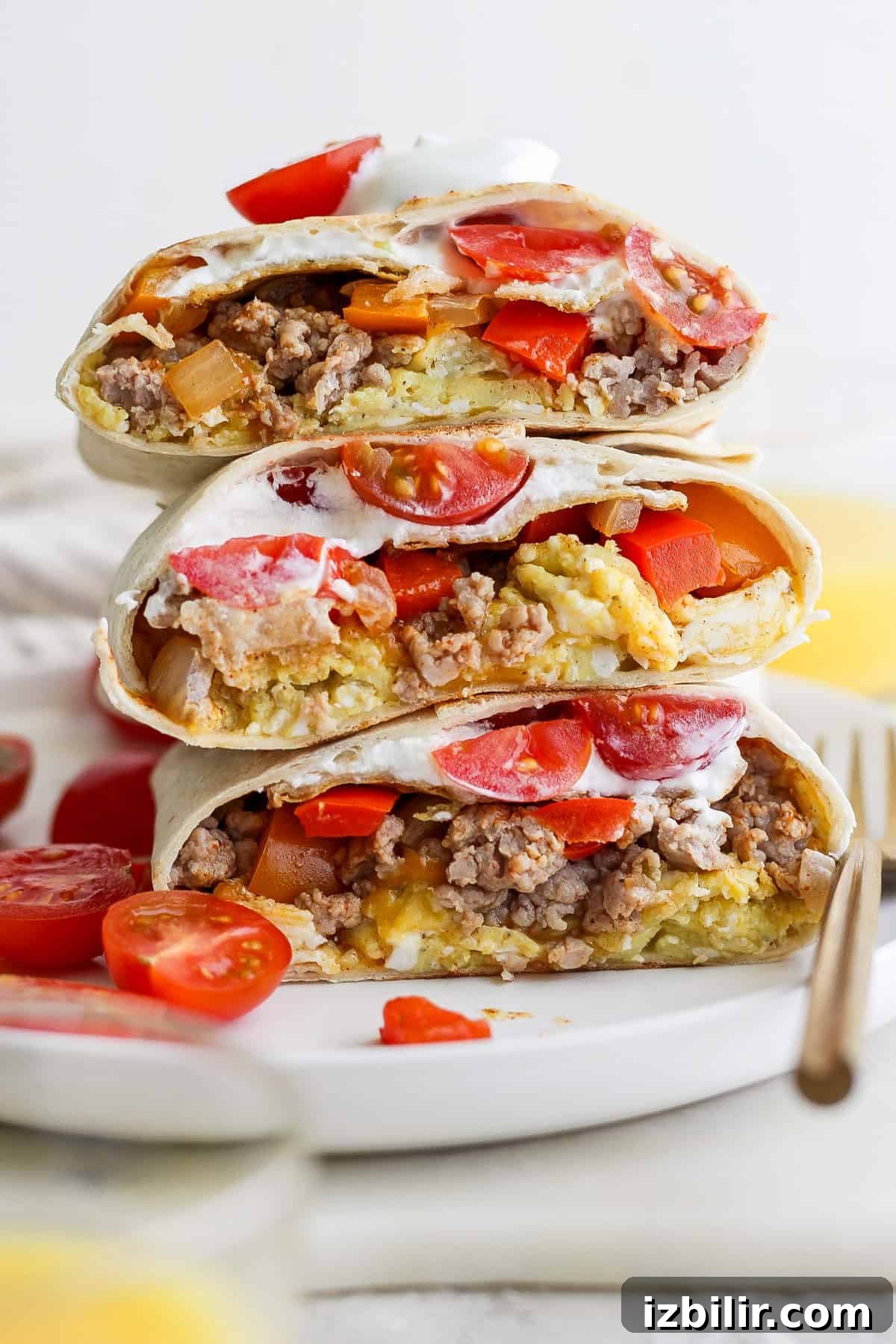 Stacked burritos filled with ground meat, cheese, tomatoes, and sour cream on a white plate. Sliced tomatoes are scattered around.