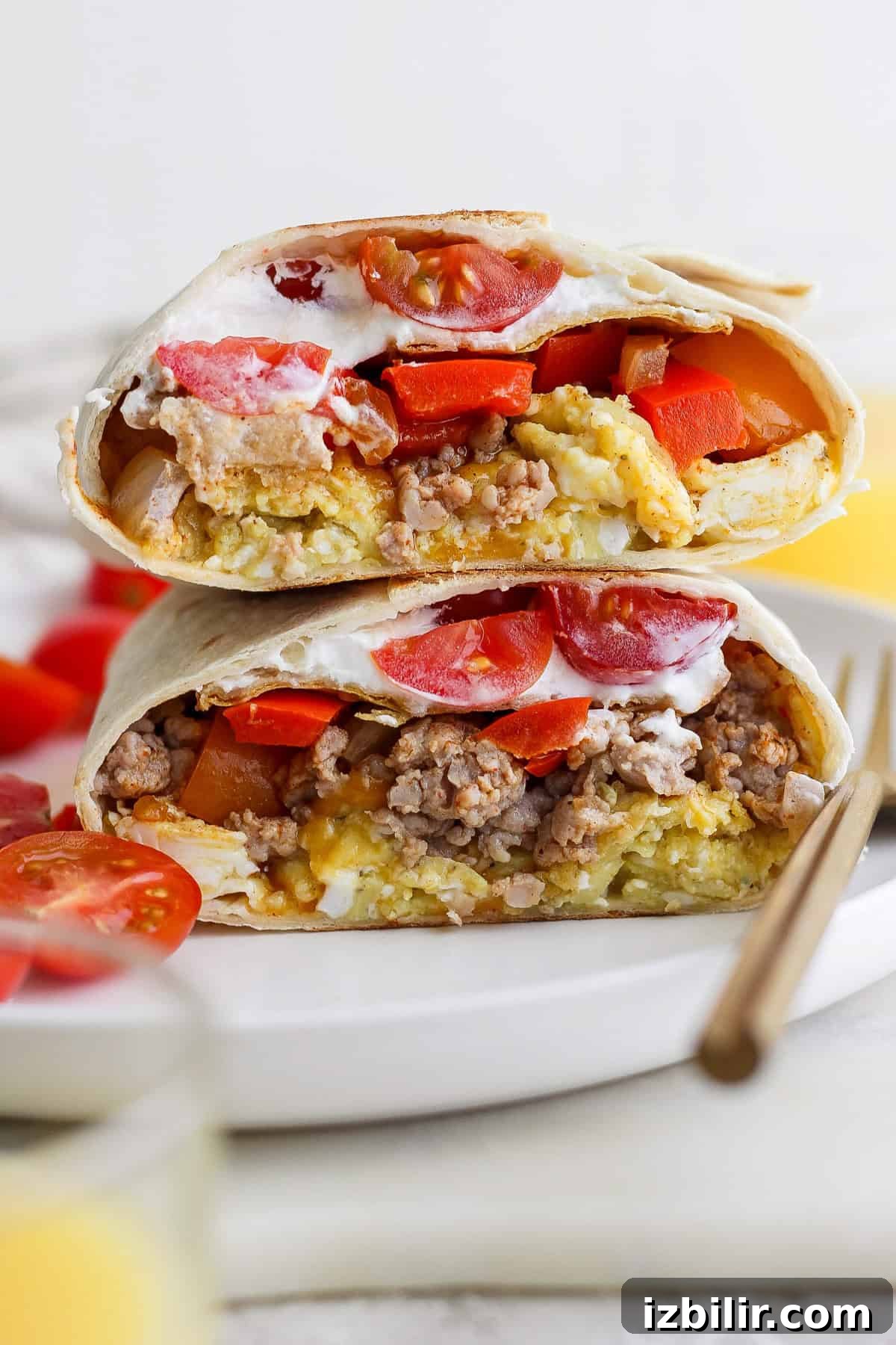 Two halves of a breakfast burrito filled with eggs, sausage, tomatoes, and topped with sour cream are stacked on a white plate.