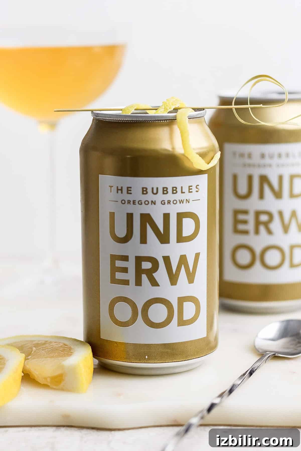 Two cans of Underwood 'The Bubbles' canned champagne cocktail with lemon garnish grace the table, accompanied by a spoon and lemon slices. A wine glass adds elegance in the background.
