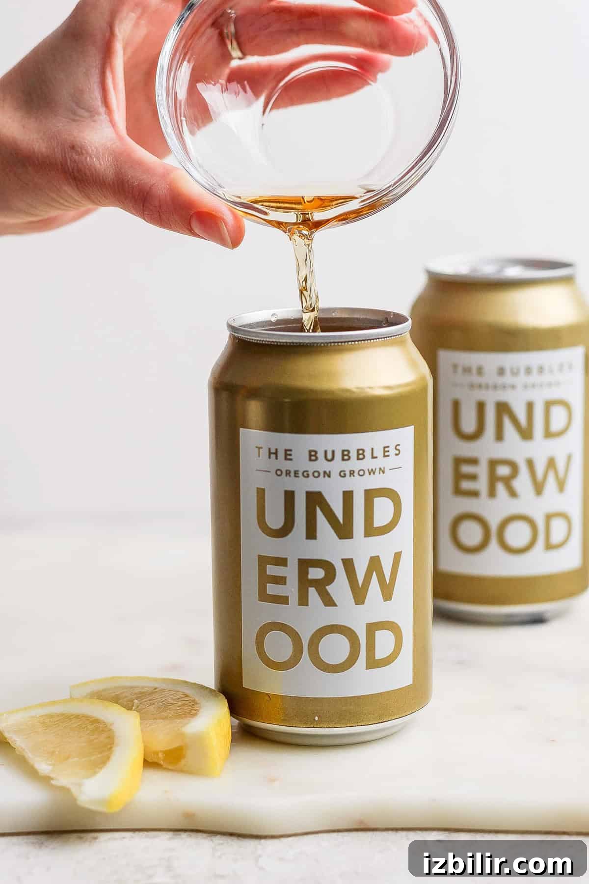 A hand elegantly pours liquid from a glass into a can labeled 'Underwood: The Bubbles Oregon Grown,' creating the perfect canned champagne cocktail. Two lemon slices and another can are subtly visible in the background, adding a touch of zest to this sparkling scene.