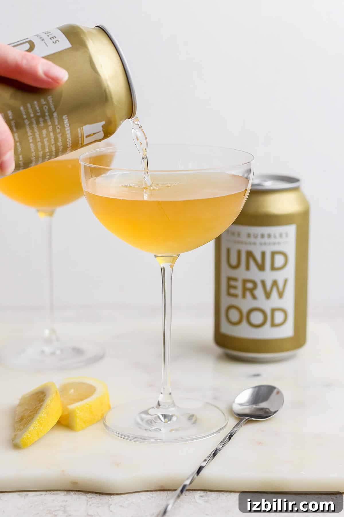 A can labeled 'Underwood' is pouring a golden liquid into a coupe glass. Another filled glass, the can itself, lemon wedges, and a spoon are arranged on the marble surface.