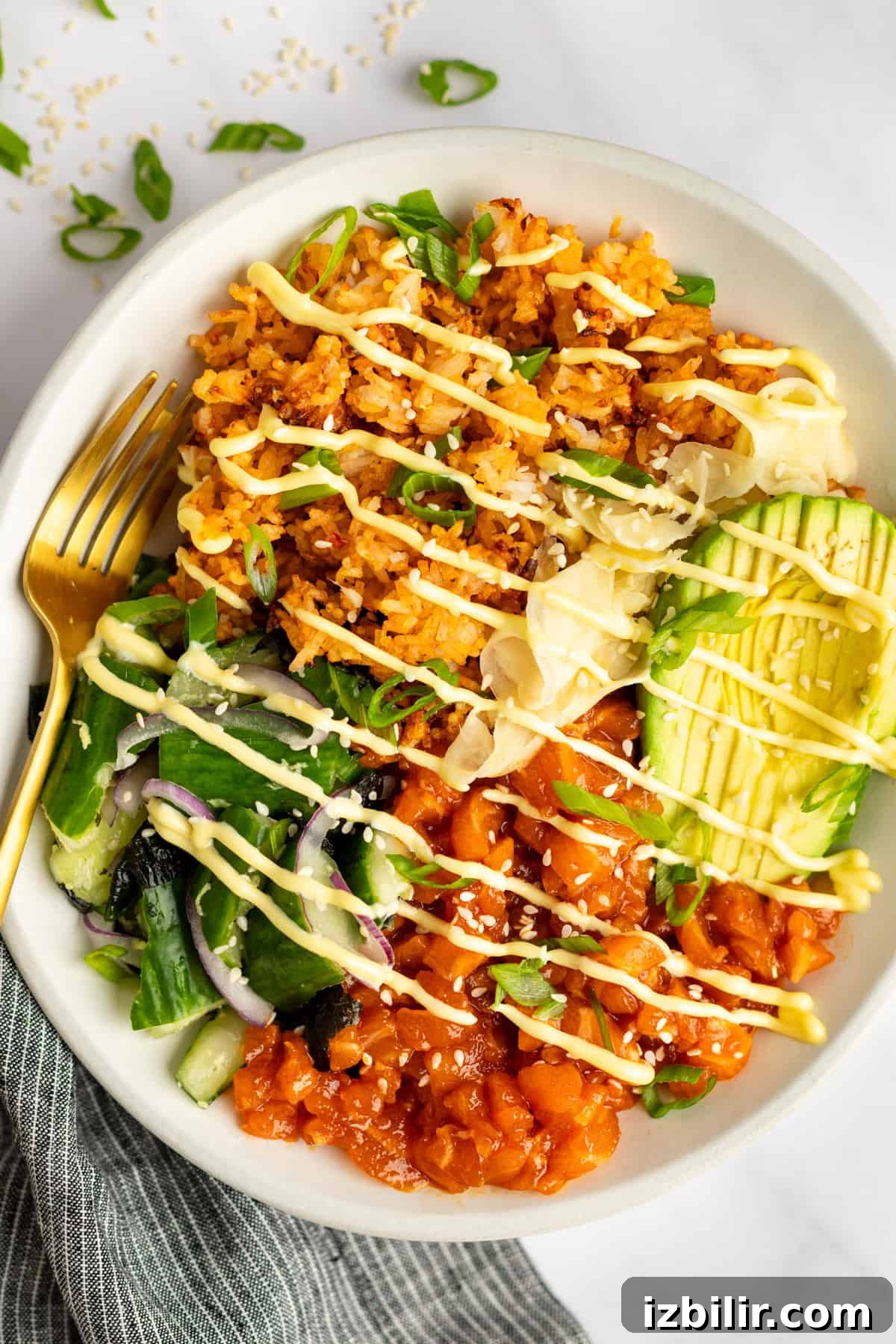 Crispy Rice Bowls with Spicy Salmon Recipe 2 A bowl of rice topped with avocado slices, marinated raw fish, sliced vegetables, shredded cabbage, and drizzled with a creamy sauce. A gold fork rests on the side.