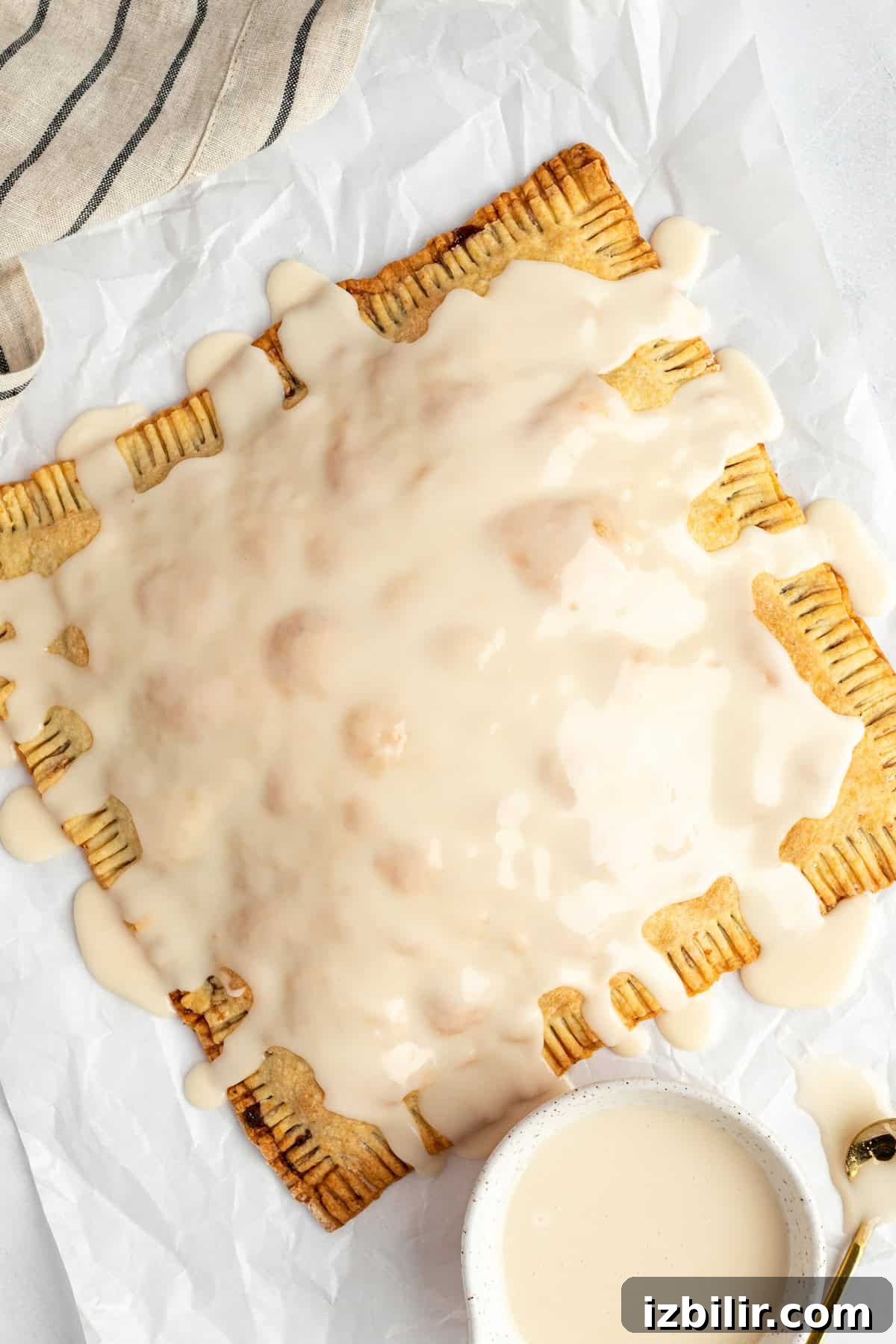 Homemade Protein Pop Tarts Recipe 6 A large rectangular pastry with ridged edges is covered in white icing, placed on crinkled parchment paper next to a small bowl of extra icing.