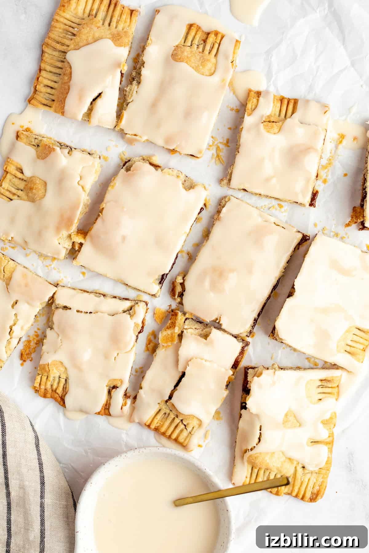 Homemade Protein Pop Tarts Recipe 7 Nine iced pastry rectangles with a crumbly texture, arranged on parchment paper. A bowl of icing with a spoon is at the bottom left.